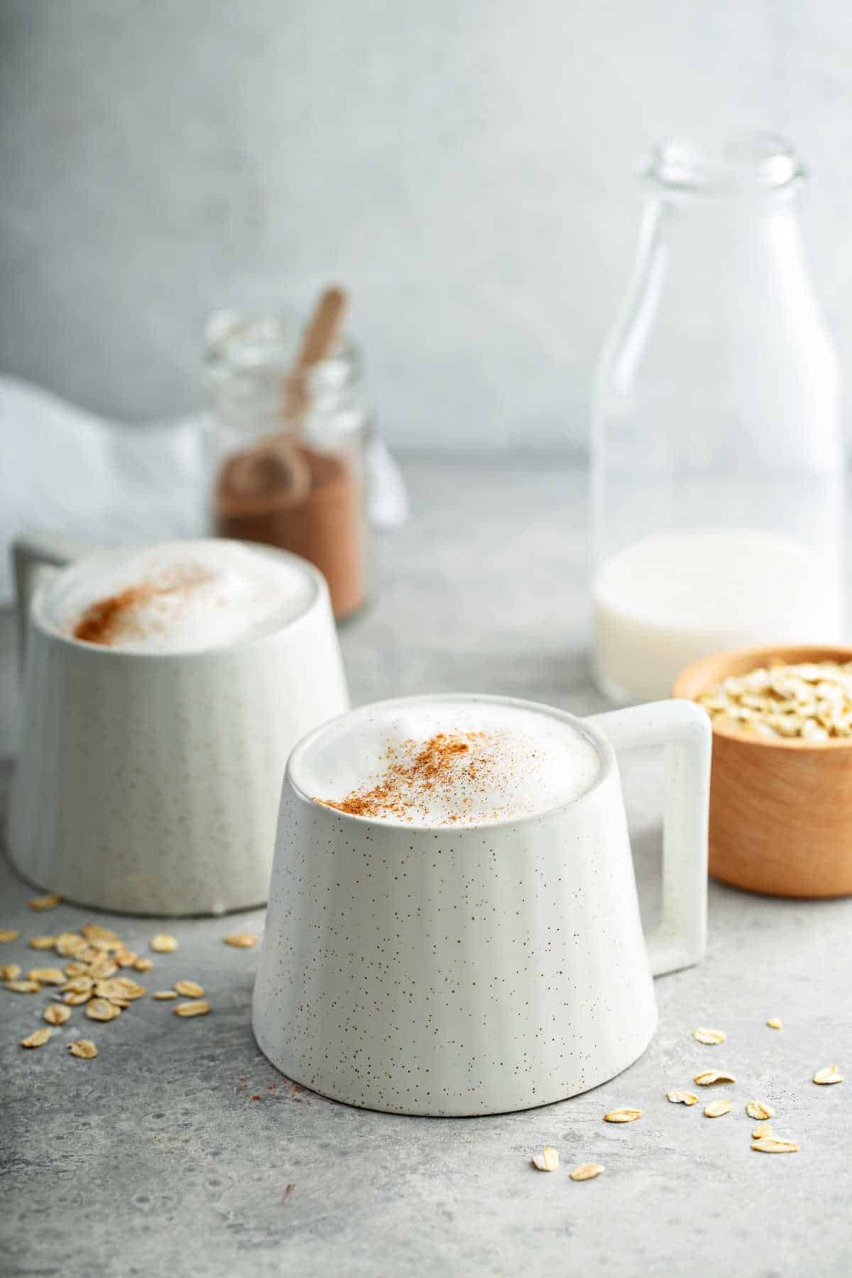 Two cups of oat milk latte, next to a jug of milk and a bowl of oats.