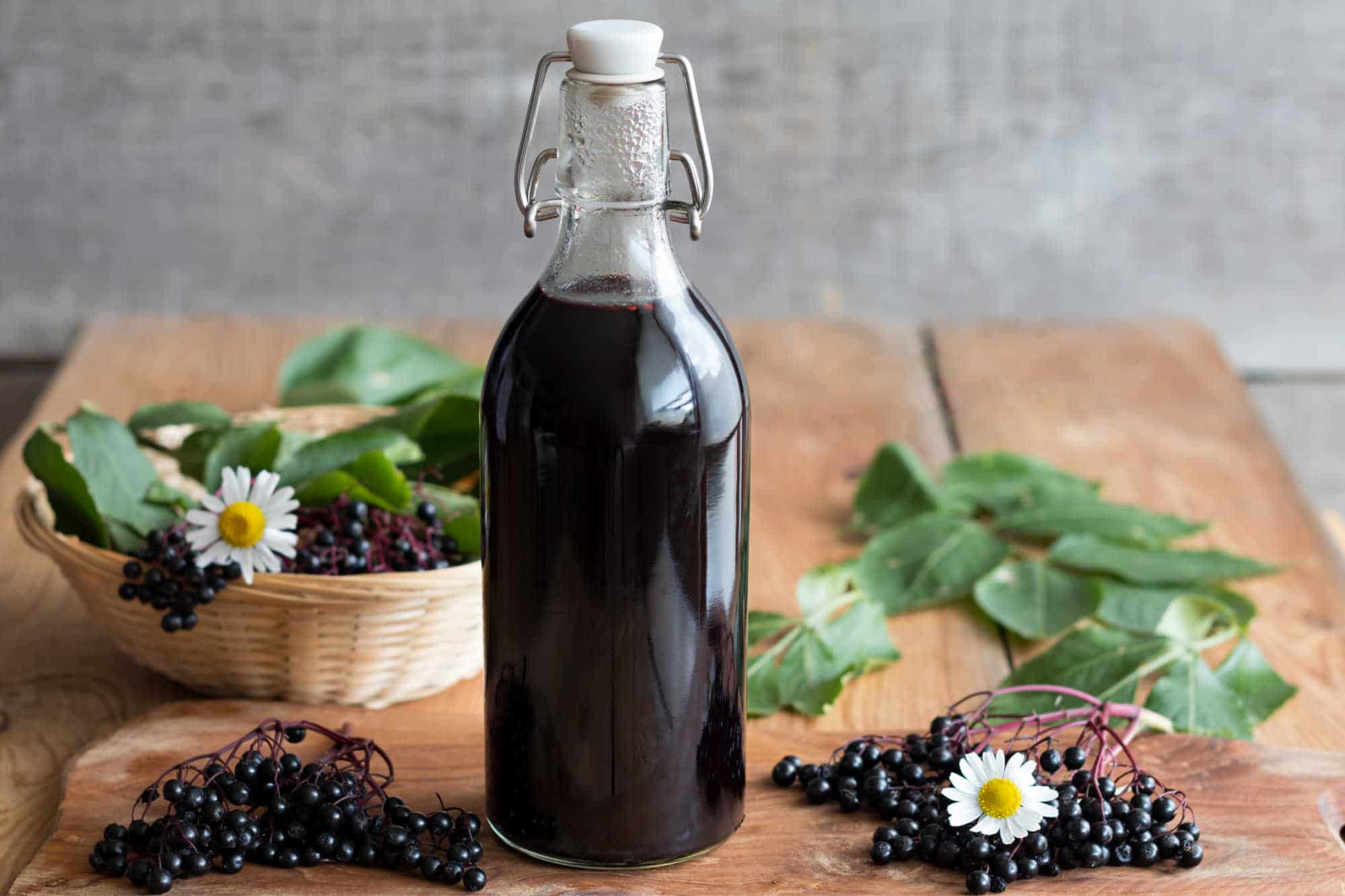 Bottle of homemade elderberry syrup, next to raw elderberries, daisies and green leaves.